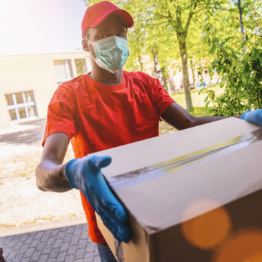 Delivery man employee in red cap blank t-shirt uniform face mask gloves hold empty cardboard box. Service quarantine pandemic coronavirus virus