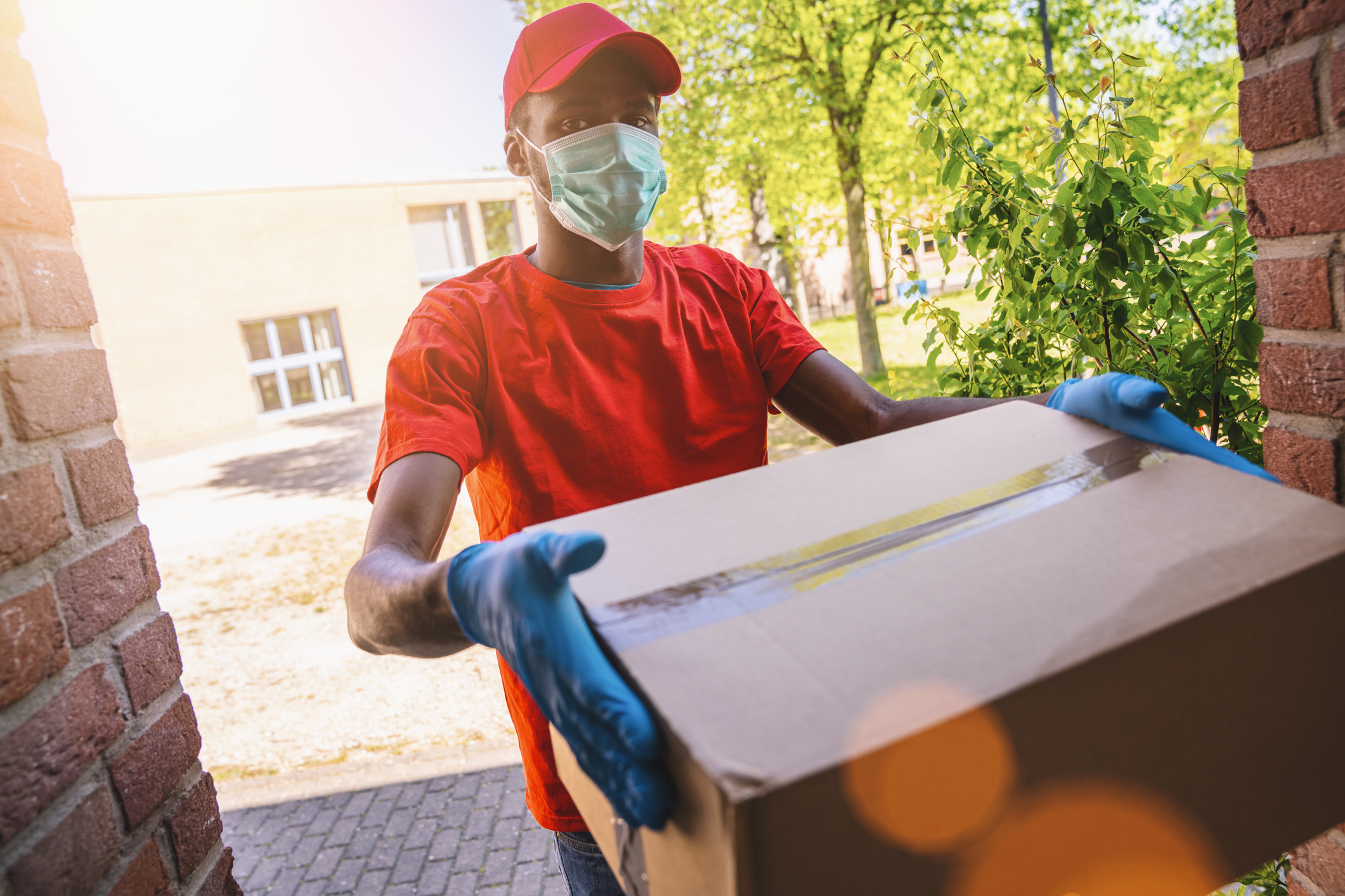 Delivery man employee in red cap blank t-shirt uniform face mask gloves hold empty cardboard box. Service quarantine pandemic coronavirus virus