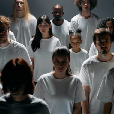 A group of people of different genders and races stand slightly apart in white t-shirts, facing the camera with blank expressions.