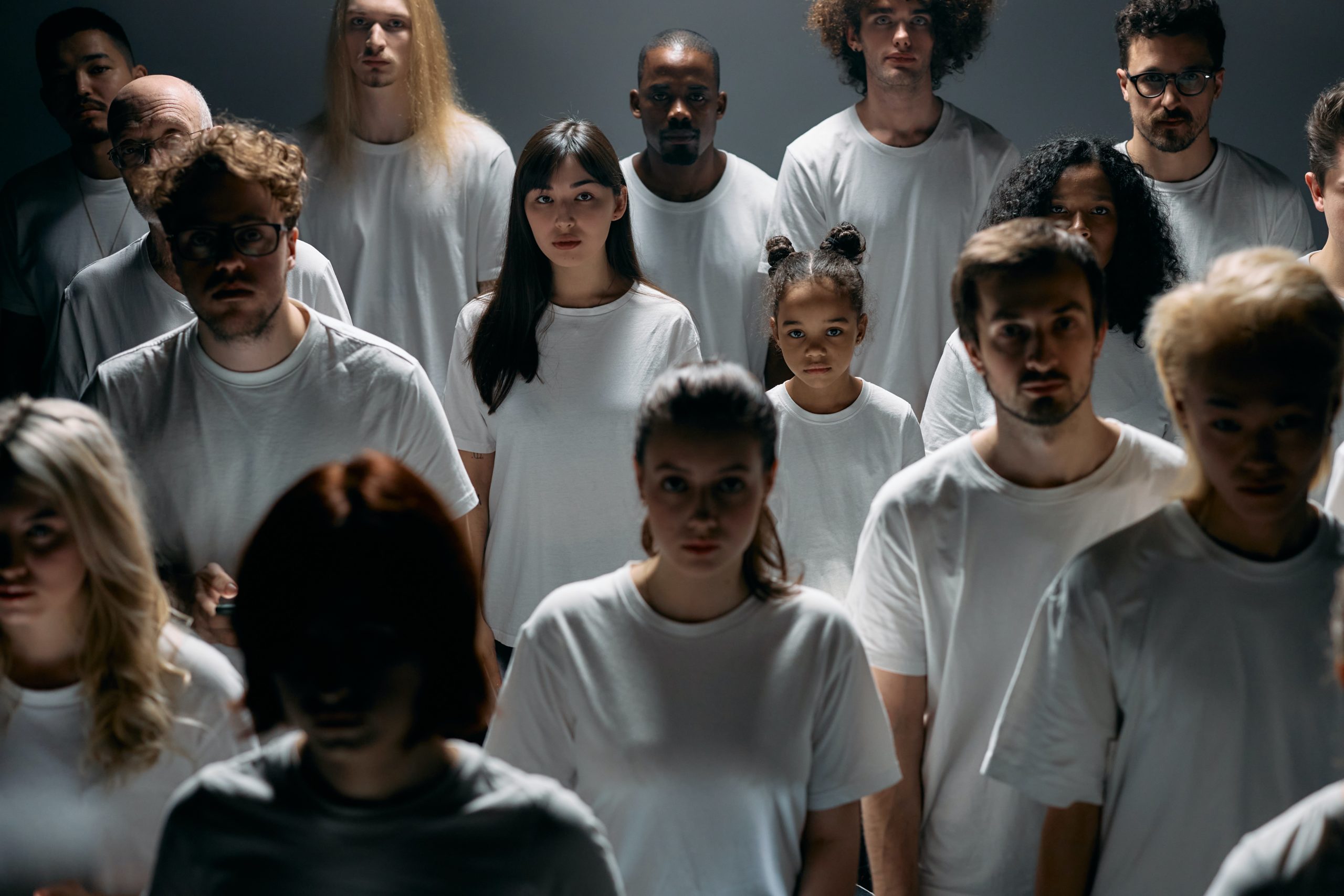 A group of people of different genders and races stand slightly apart in white t-shirts, facing the camera with blank expressions.