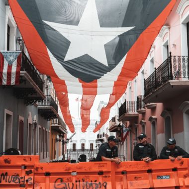 Old San Juan, Puerto Rico: two sets of apartments face each other across a street divided a giant Puerto Rican flag that stretches above them, in front the street is blocked off by police