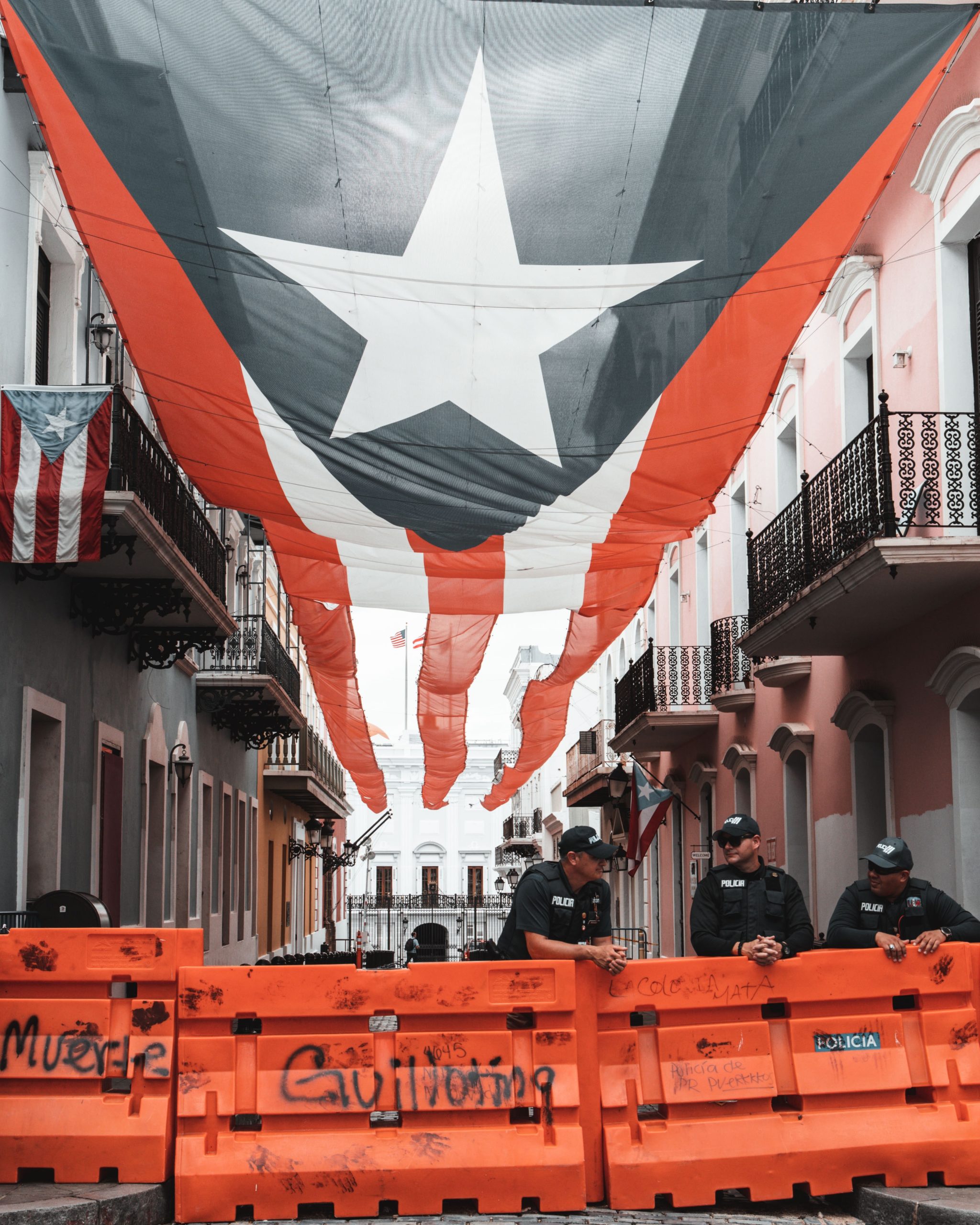 Old San Juan, Puerto Rico: two sets of apartments face each other across a street divided a giant Puerto Rican flag that stretches above them, in front the street is blocked off by police